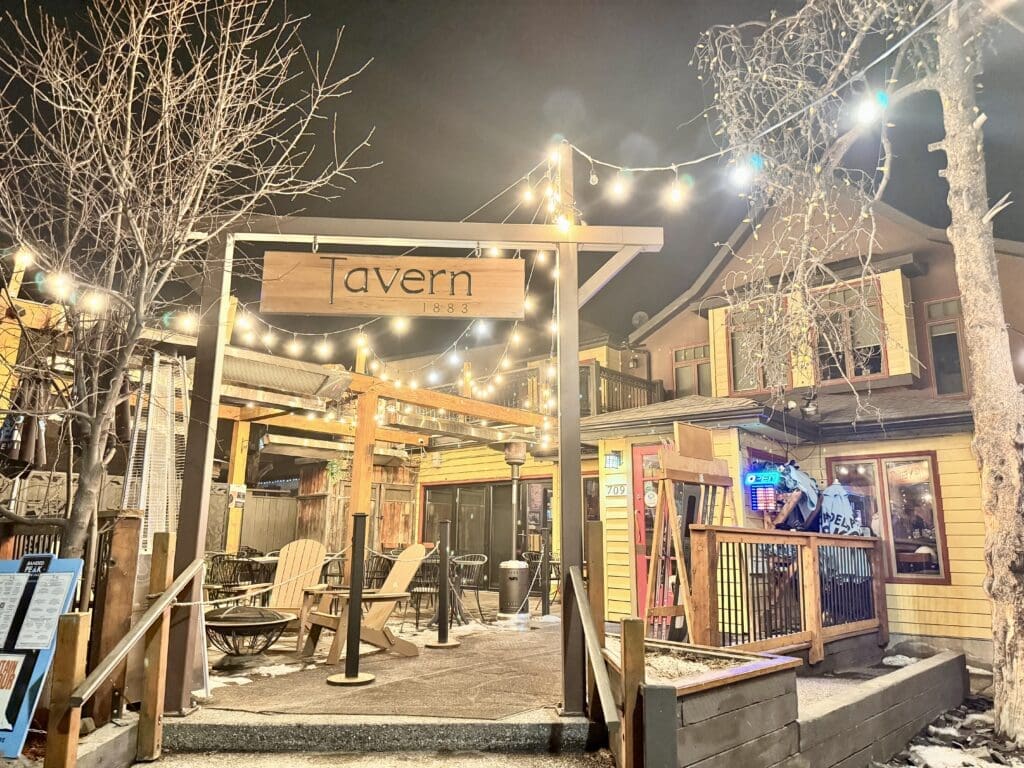 Cozy outdoor patio at Tavern 1883 in Canmore, Alberta, illuminated with string lights on a winter night. The rustic restaurant features Adirondack chairs, wooden decor, and a welcoming atmosphere perfect for evening dining and drinks in the Canadian Rockies.