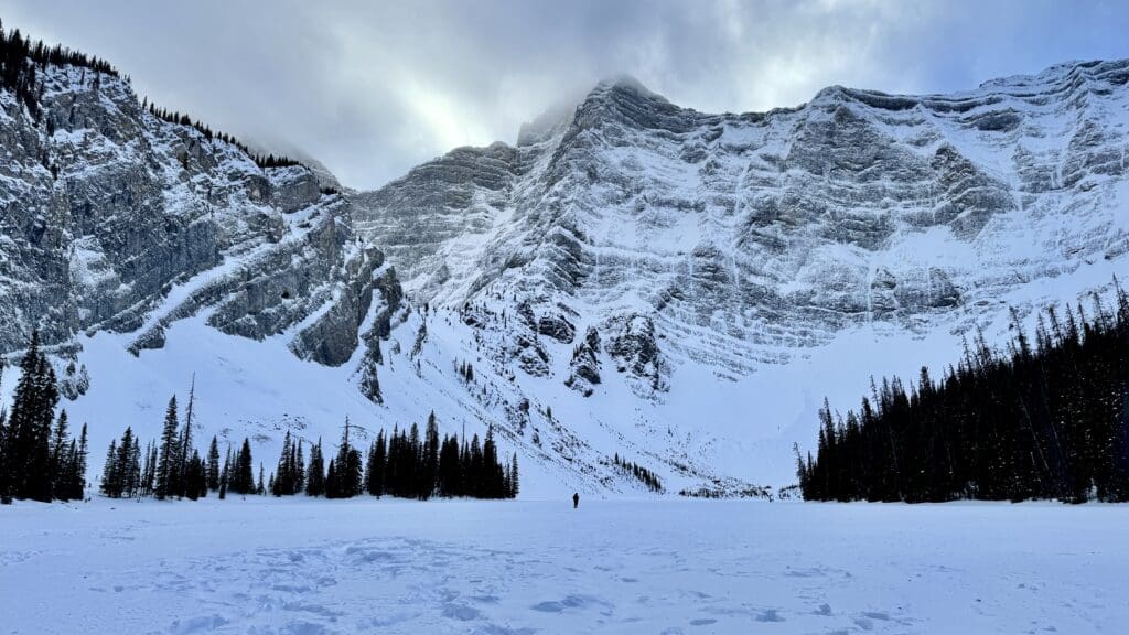 A solitary figure walks across a snowy Rawson Lake, one of the best hikes in Kananaskis, beneath imposing, snow-covered Mount Sarrail. Tall pine trees line the foreground, under a cloudy sky.