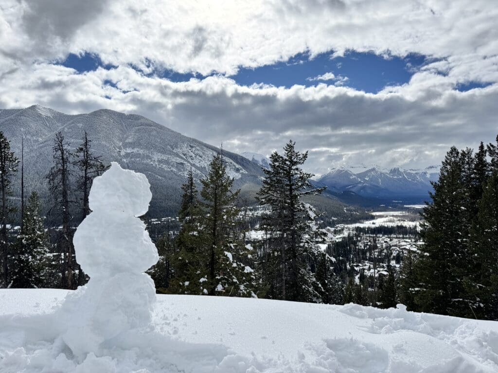 A small snowman perched on Tunnel Mountain Trail gazes out over the snowy town of Banff, with a stunning backdrop of snow-covered peaks and dramatic winter skies.