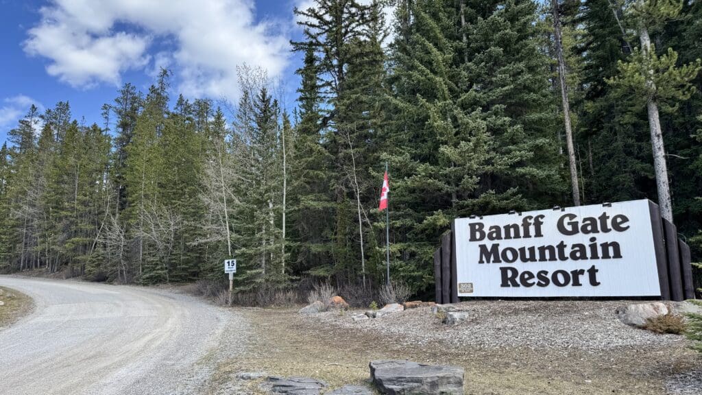 Entrance sign for Banff Gate Mountain Resort surrounded by dense pine forest and a winding gravel road, with a Canadian flag waving under a partly cloudy sky.