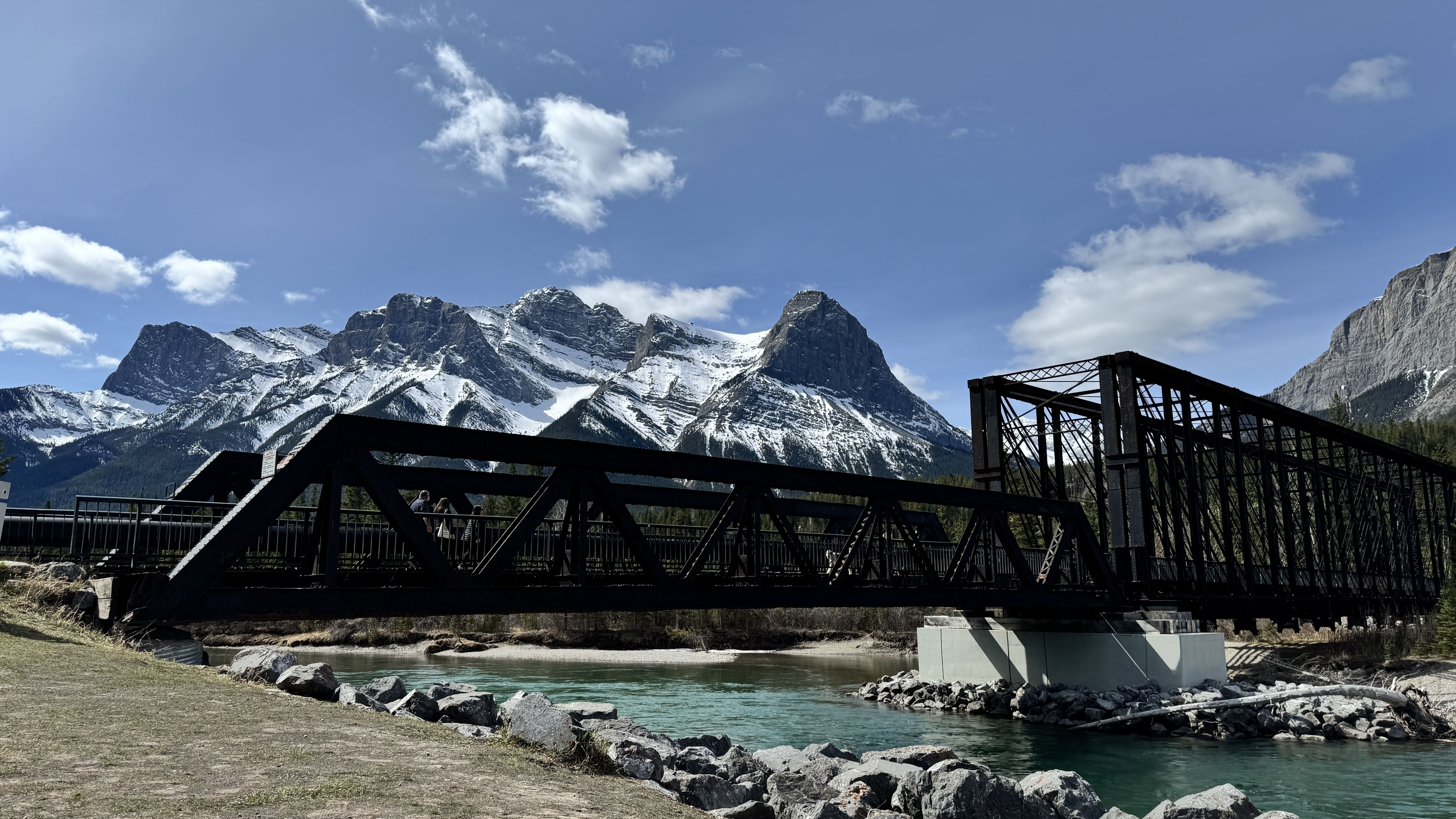Historic Engine Bridge spanning the turquoise Bow River in Canmore, Alberta, with a backdrop of the snow-capped Rocky Mountains under a clear blue sky.