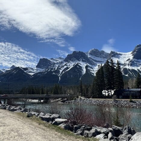 Scenic walking path along Bow River in Canmore, Alberta, featuring Canmore’s Bridge Road crossing the river, with towering Rocky Mountains in the background under a bright blue sky.