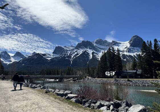 Scenic walking path along Bow River in Canmore, Alberta, featuring Canmore’s Bridge Road crossing the river, with towering Rocky Mountains in the background under a bright blue sky.