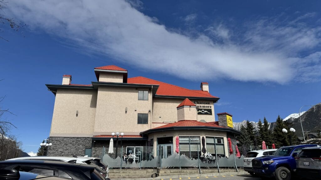 Exterior view of Chez Francois Restaurant and Patio at Chateau Canmore, featuring a beige and stone building with red roofs, outdoor seating with mountain-themed frosted glass dividers, and a backdrop of the Canadian Rockies under a vibrant blue sky. The best Restaurants to Dine in Canmore