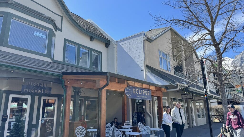 Front view of Eclipse Coffee Roasters in downtown Canmore, nestled in a cozy building with outdoor seating, wooden patio accents, and a scenic mountain backdrop under a clear blue sky. The best Restaurants to Dine in Canmore