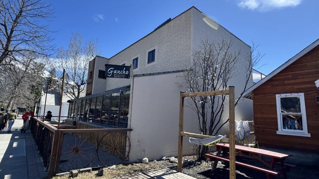 Exterior view of Gaucho Brazilian Barbecue restaurant in Canmore, Alberta, featuring a rustic patio with wagon wheel decor and a white brick facade under a clear blue sky.