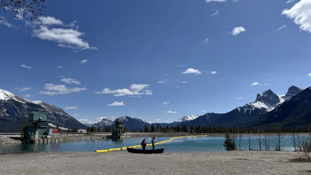 Two people preparing a canoe on the shoreline of Rundle Forebay Reservoir in Canmore, Alberta, with the stunning backdrop of the Three Sisters mountains under a clear blue sky.
