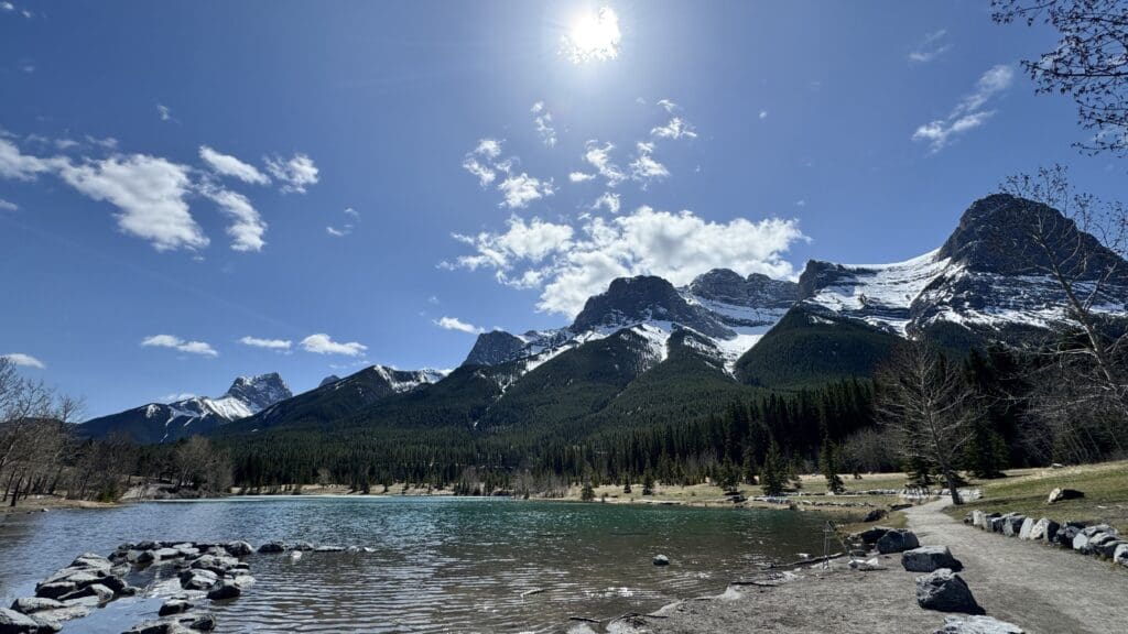 Scenic view of Quarry Lake in Canmore, Alberta, featuring crystal-clear water, rocky shoreline, a forested backdrop, and the majestic snow-capped Rocky Mountains under a bright blue sky.