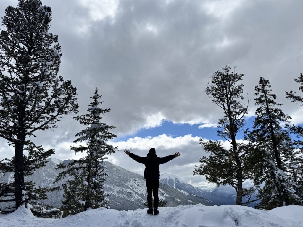 Silhouette of a hiker standing triumphantly at the snow-covered summit of Tunnel Mountain in Banff, arms outstretched against a dramatic mountain backdrop and moody skies.