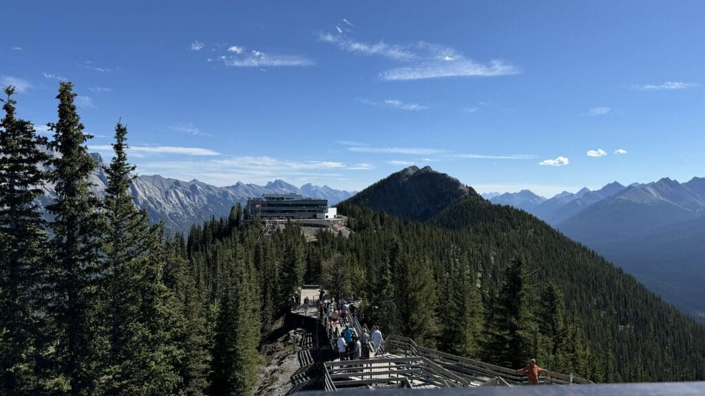 Many people walk on the wide boardwalk from Sky Bistro to Cosmic Ray Station in Sulphur Mountain in Banff National Park with lots on mountain at the back