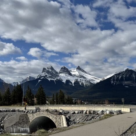 Scenic view of the iconic Three Sisters peaks from the Cougar Creek Trailhead in Canmore, Alberta, with a pedestrian bridge and cloudy sky enhancing the dramatic mountain backdrop.
