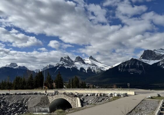Scenic view of the iconic Three Sisters peaks from the Cougar Creek Trailhead in Canmore, Alberta, with a pedestrian bridge and cloudy sky enhancing the dramatic mountain backdrop.