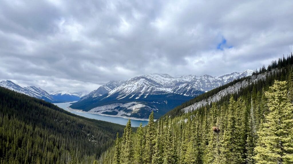Vast mountain range including Goat Mountain in Kananaskis, Alberta with snow-capped peaks, dense pine forest, and a serene blue lake under a cloudy sky. Majestic and tranquil landscape.