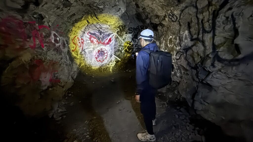 A person with a backpack and cap shines a flashlight on colorful graffiti on a dark cave wall inside Heart Creek Bunker in Kananaskis, Alberta. The scene feels mysterious and adventurous.