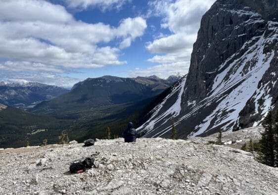 Person sits on rocky mountaintop of West Wind Pass in Kananaskis, Alberta, facing expansive mountain range under cloudy blue sky. Snow patches on steep cliff; serene, contemplative mood.