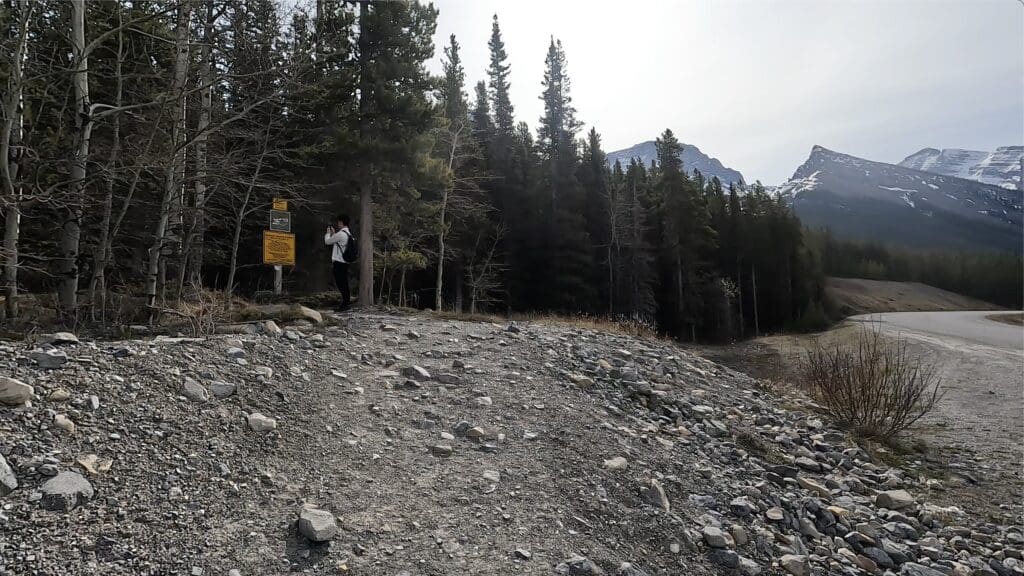 A person with a backpack photographing a forest entrance of West Wind Pass in Kananaskis, Alberta with warning signs. Rocky terrain and tall pine trees with snow-capped mountains in the background.