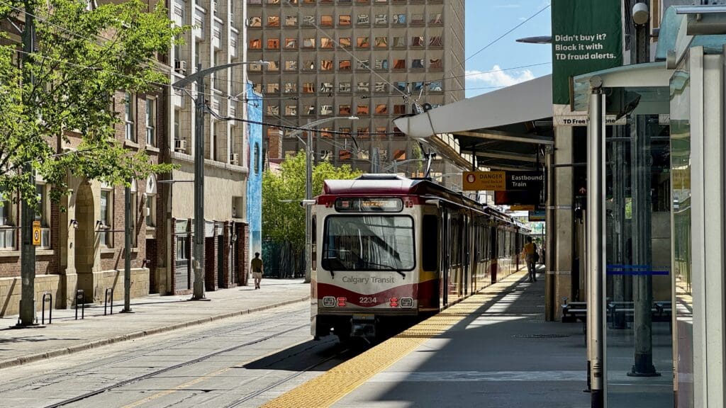A Calgary Transit train in Alberta arrives at a sunny city street station. Tall buildings line the street, casting shadows. A person walks on the platform.