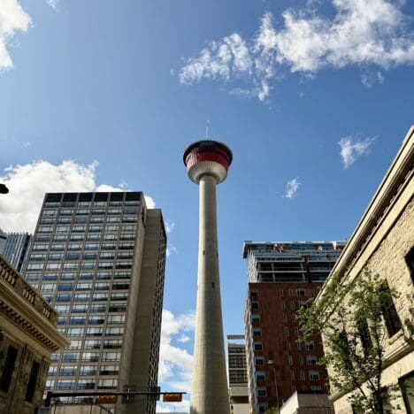 Tall Calgary Tower in Alberta under a blue sky with scattered clouds, surrounded by urban buildings. Cars and trees line the street, creating a vibrant cityscape.