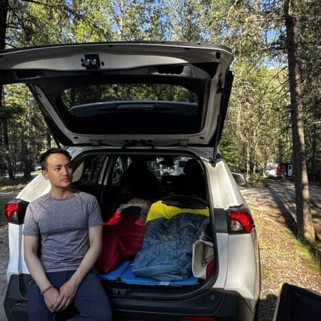 A man sits on the back of an SUV, open trunk filled with colorful sleeping bags, surrounded by a sunlit forest at Redstreak Campground in Kootenay National Park, BC.