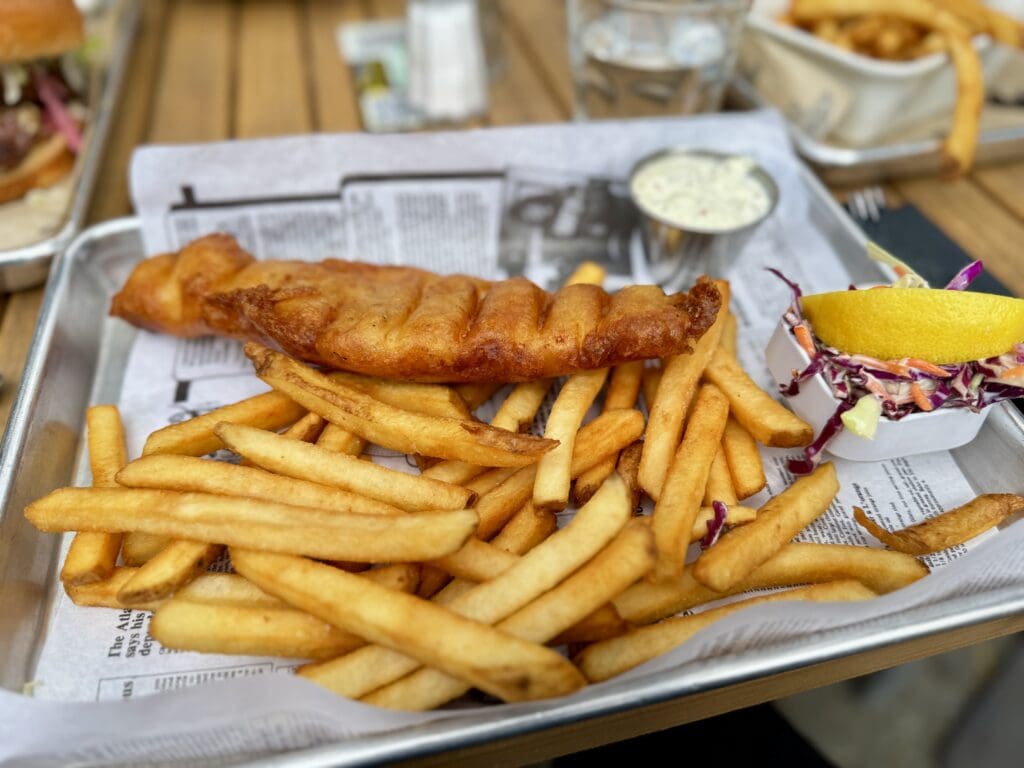 A tray of golden-brown fish and chips served with coleslaw and a lemon wedge, accompanied by tartar sauce. The presentation is casual yet appetizing.