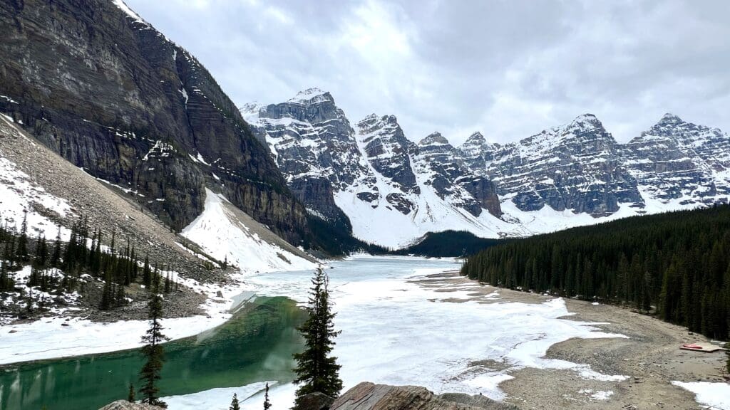 Snow-capped Valley of the Ten Peaks surround a partially frozen turquoise Moraine Lake in banff National Park, with evergreen trees lining the shoreline. The scene is serene and majestic.