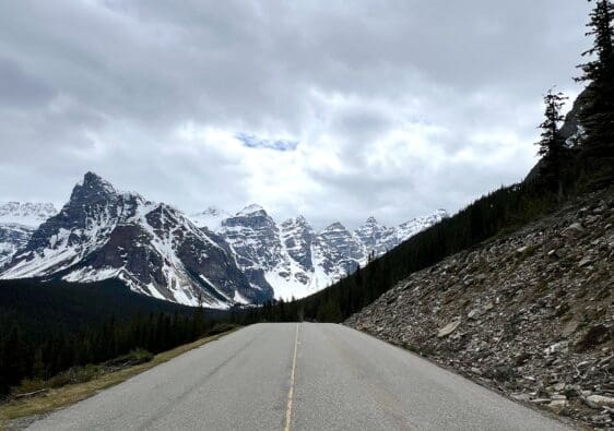 The deserted Moraine Lake Road stretches toward snow-capped mountains under a cloudy sky. Pine trees line the road, creating a serene and majestic landscape.