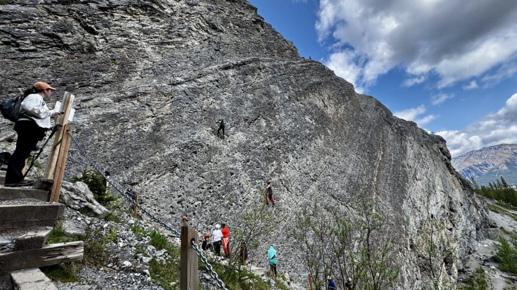 A group of climbers scale a steep rocky cliff under a partly cloudy blue sky. A person observes from wooden steps, surrounded by lush greenery. Energetic and adventurous.
