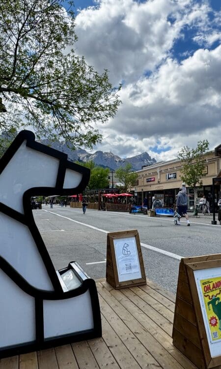 Outdoor street scene in Canmore Town, Alberta with a geometric polar bear sculpture on a wooden platform. People stroll past rustic buildings under a cloudy sky, with mountain peaks in the background.