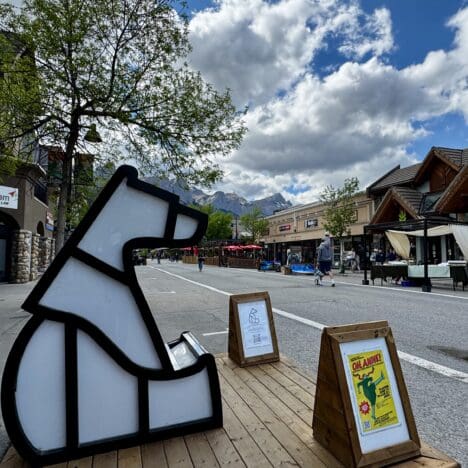 Outdoor street scene in Canmore Town, Alberta with a geometric polar bear sculpture on a wooden platform. People stroll past rustic buildings under a cloudy sky, with mountain peaks in the background.