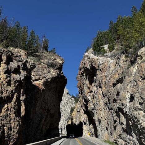 A car drives through the narrow Sinclair Canyon in Kootenay National Park, BC with towering, rugged rock walls under a clear blue sky. Tall trees line the cliff edges, adding natural beauty.