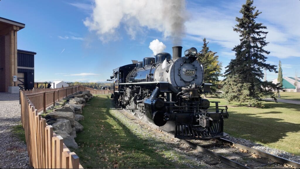 A black steam locomotive numbered 2023 at Heritage in Calgary, Alberta emits white smoke under a vibrant blue sky. Surrounded by trees, it runs past a wooden fence and a brick building, conveying nostalgia.