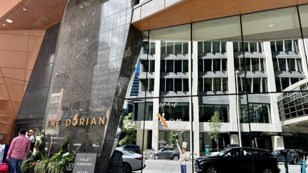 The image shows the entrance of The Dorian Hotel in Calgary, Alberta with a reflective glass facade. Two people stand nearby. The tone is modern and sophisticated.