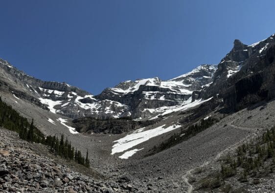 Snow-dusted Stanley Glacier in Kootenay National Park, BC under a clear blue sky, with rugged rocky terrain and scattered evergreen trees in the foreground, evoke a serene, majestic feel.