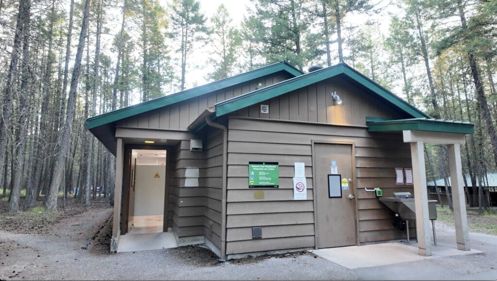 A brown wooden restroom building in a forest setting at Redstreak Campground in Radium, BC, with a green roof. Signs are visible and a path leads into the surrounding tall trees.