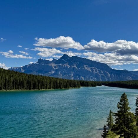 Scenic view of Two Jack Lake in Banff National Park, Alberta surrounded by dense evergreen forests, with majestic Mount Rundle under a clear blue sky and scattered clouds. Peaceful and serene.