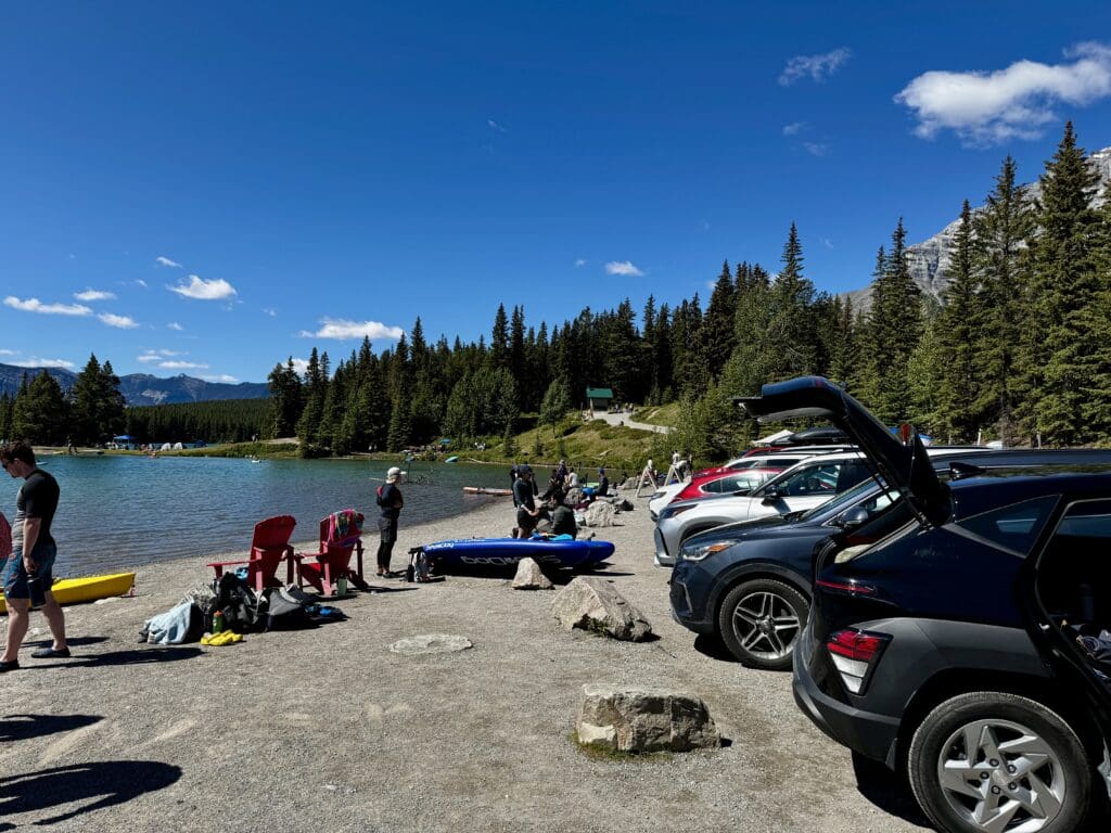 A scenic lakeside parking area in Two Jack Lake in Banff National Park, Alberta with pine trees and a mountain backdrop. People in swimsuits enjoy the sunny day, with cars lined up on the gravel.