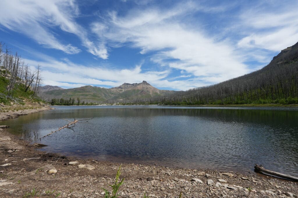 Crandell Lake in Waterton Lakes National Park, Alberta with clear blue sky and wispy clouds. The calm water reflects surrounding green hills and burned trees, creating a tranquil scene.