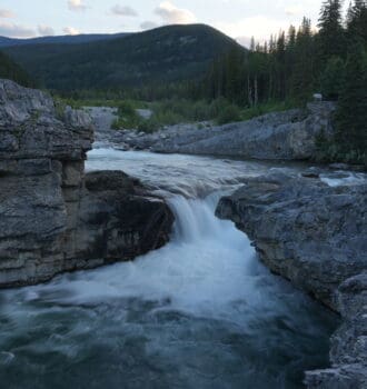 Rocky waterfall in Elbow Falls, Alberta scene with rushing water cascading between large gray boulders. Lush pine forest and distant mountains under a cloudy sky at dusk.