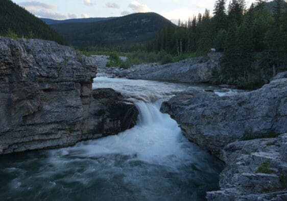Rocky waterfall in Elbow Falls, Alberta scene with rushing water cascading between large gray boulders. Lush pine forest and distant mountains under a cloudy sky at dusk.