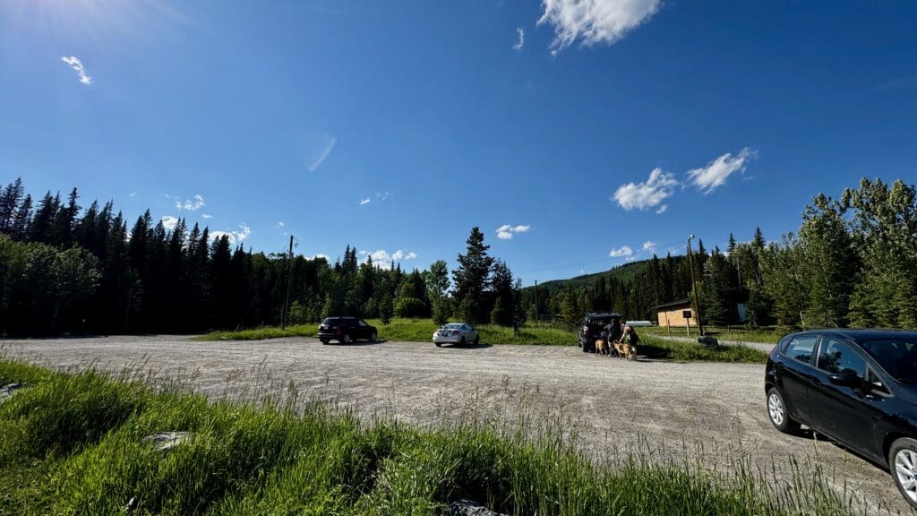 A serene parking area of Fullerton Loop Trailhead in Kananaskis, Alberta bordered by lush pine trees with a clear blue sky. Several parked cars, people with dogs, and a wooden cabin create a peaceful, sunny day.