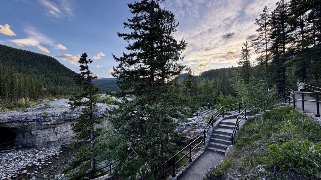 Scenic sunset view of a forested landscape with a path and railing besides Elbow Falls in Kanananaskis, Alberta. A bridge spans a rocky riverbed. The sky is lit with warm hues of orange and pink.