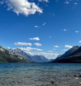 Waterton Lake, Alberta with clear, rippling water reflects towering mountains under a bright blue sky with scattered clouds. A person sits on the rocky shore.