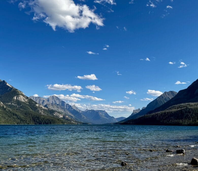 Waterton Lake, Alberta with clear, rippling water reflects towering mountains under a bright blue sky with scattered clouds. A person sits on the rocky shore.