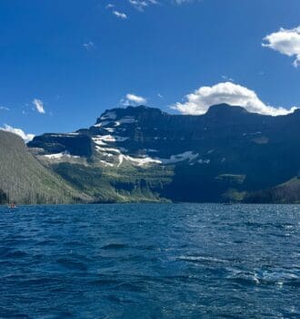 A serene mountain Cameron Lake in Waterton Lakes National Park, Alberta with rippling blue water under a clear sky. Snow-capped peaks rise in the background, evoking tranquility and grandeur.