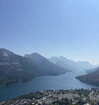 A serene landscape of Waterton Lake in Waterton Lakes National Park, Alberta surrounded by mountains under a clear sky. In the foreground, there is a small town with scattered buildings.