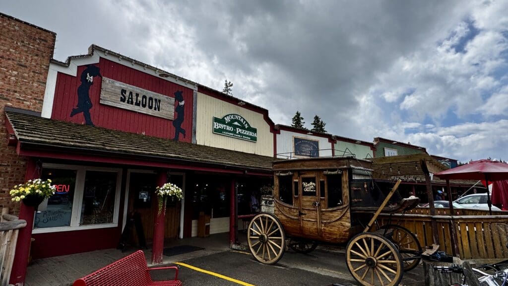 Western-style storefronts in Bragg Creek, Alberta, including a saloon and bistro, under cloudy skies. A vintage wooden carriage is parked in front, creating a nostalgic atmosphere.